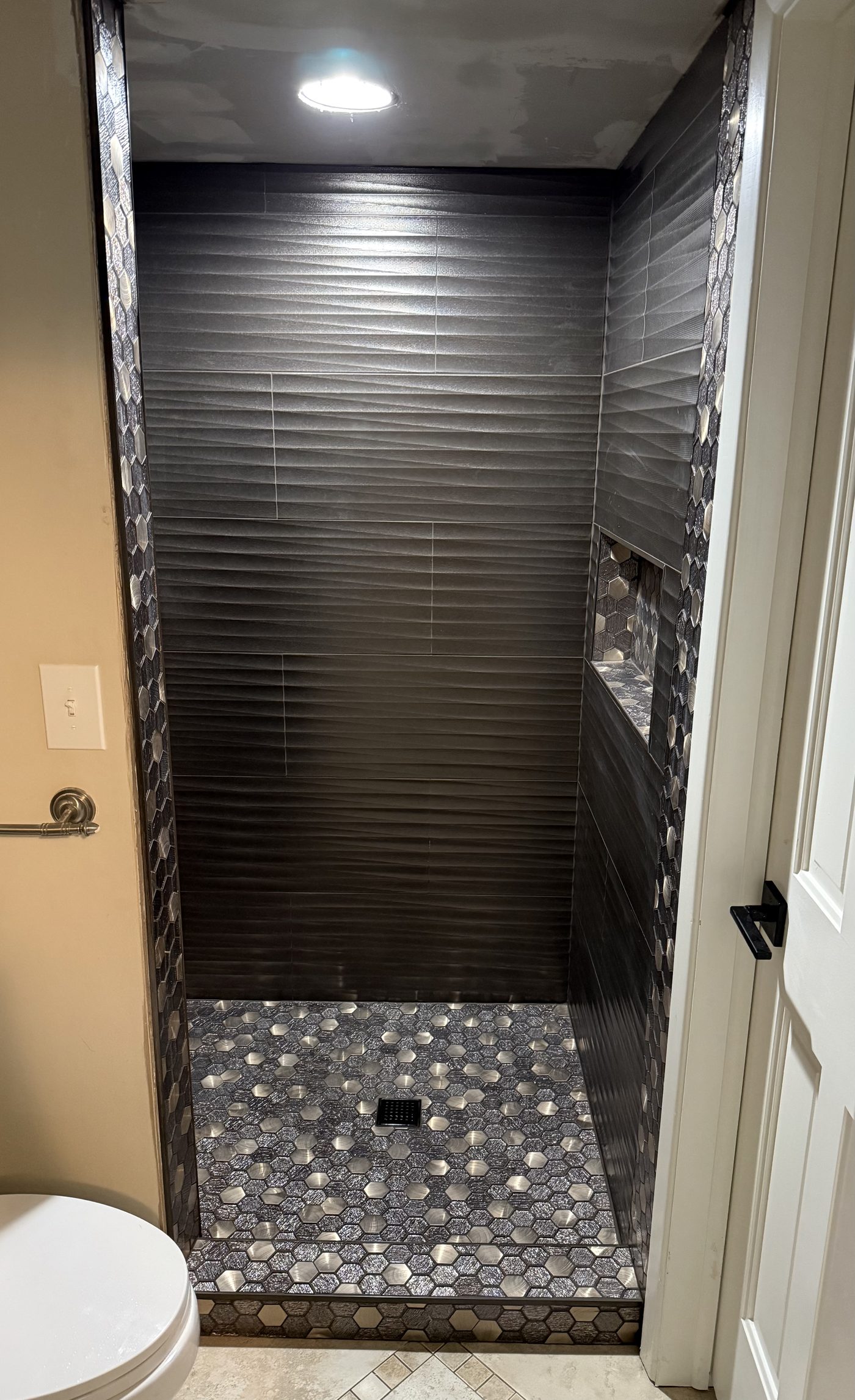 Bold walk-in shower with dark charcoal ribbed horizontal tile walls and hexagon metal mosaic floor in Lake County Ohio
