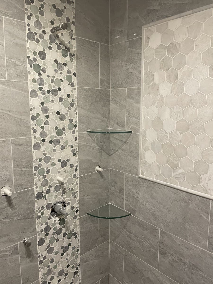 Walk-in shower with gray stone large-format tile, pebble mosaic accent strip, hexagon marble niche, and glass corner shelves in Lake County Ohio