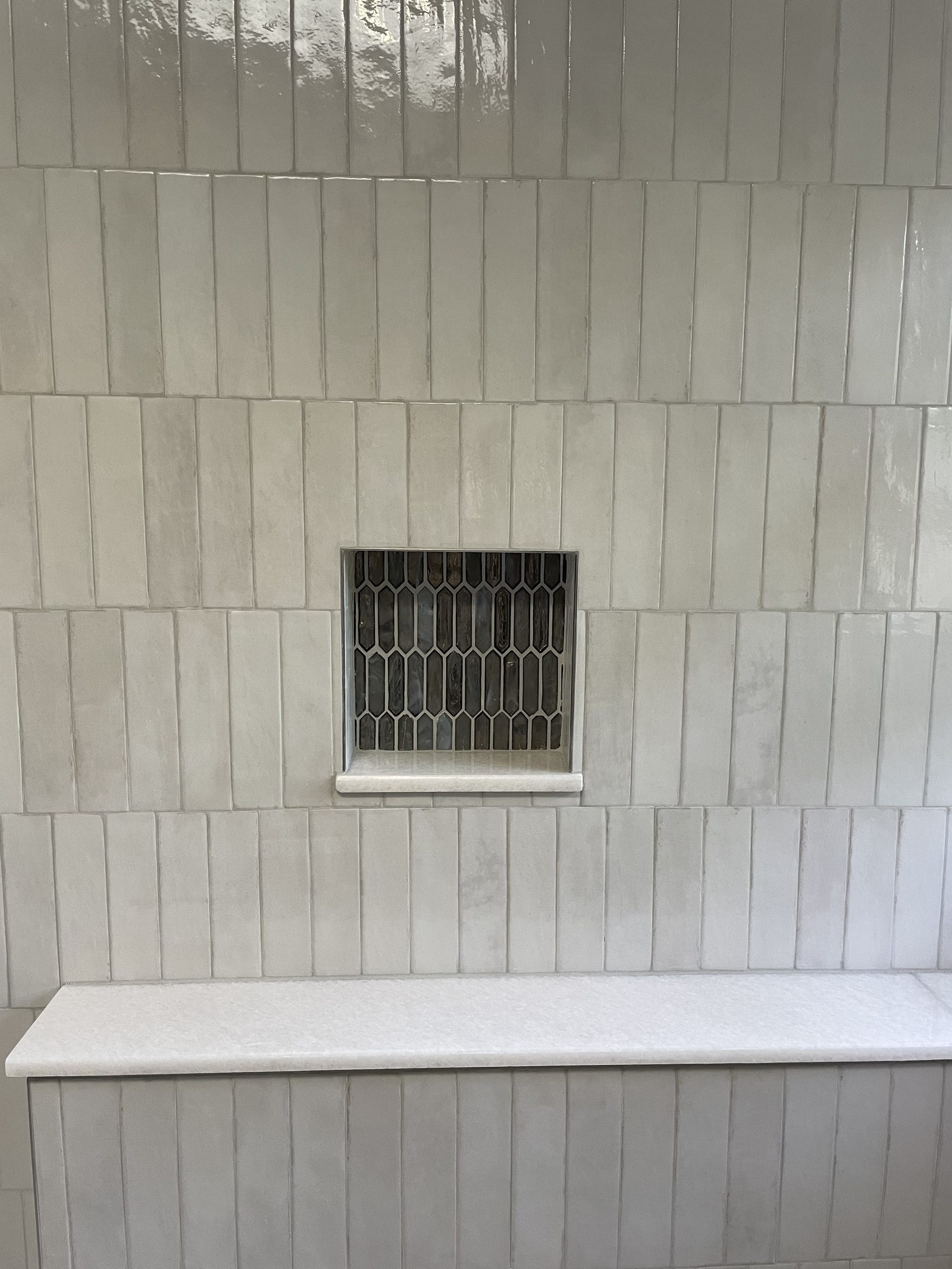 Shower wall detail with vertical white zellige stick tile and custom dark gold picket mosaic niche with glass shelf in Lake County Ohio