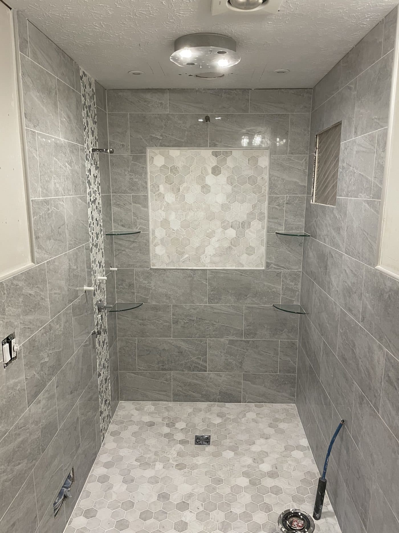 Large walk-in shower with grey stone tile, pebble mosaic accent strip, hexagon marble niche, and glass corner shelves in Northeast Ohio