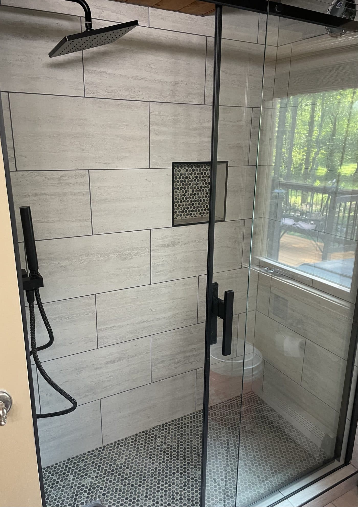 Walk-in shower with large-format grey tile, matte black rain head, penny round mosaic niche, and frameless black frame glass door in Northeast Ohio