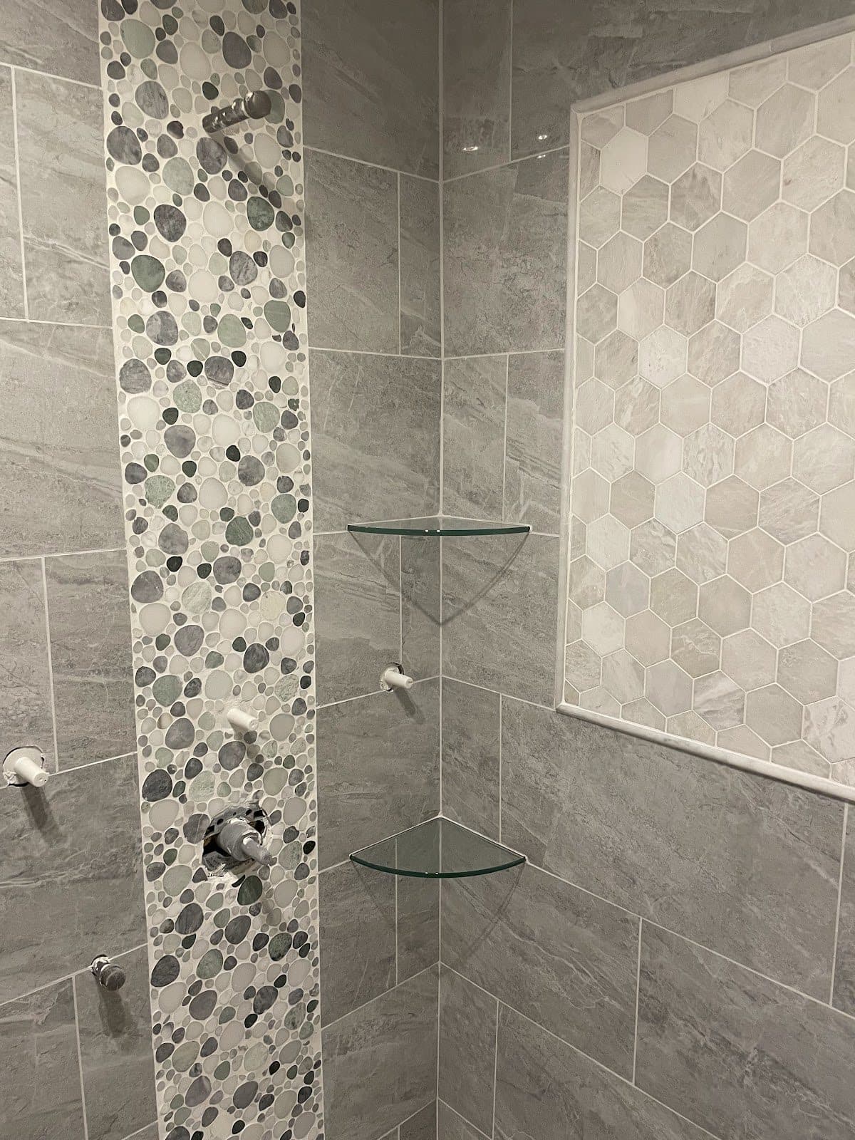 Walk-in shower with gray stone tile, colorful pebble mosaic accent strip, hexagon marble niche, and glass corner shelves in Cuyahoga County